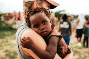 A white woman holding a black toddler in Haiti. There are peopel in the background. The baby needed a hug it seems.