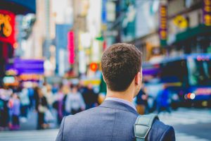 Business person in a suit carrying a laptop bag and heading twowrad city centre with lots of people and ads