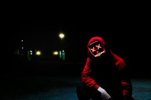 Creepy looking guy wearing a red hoody squats in the dark. His eyes are ligh graffiti crosses, his mouth a light line. It seems there is a car in the background - we see the headlights.